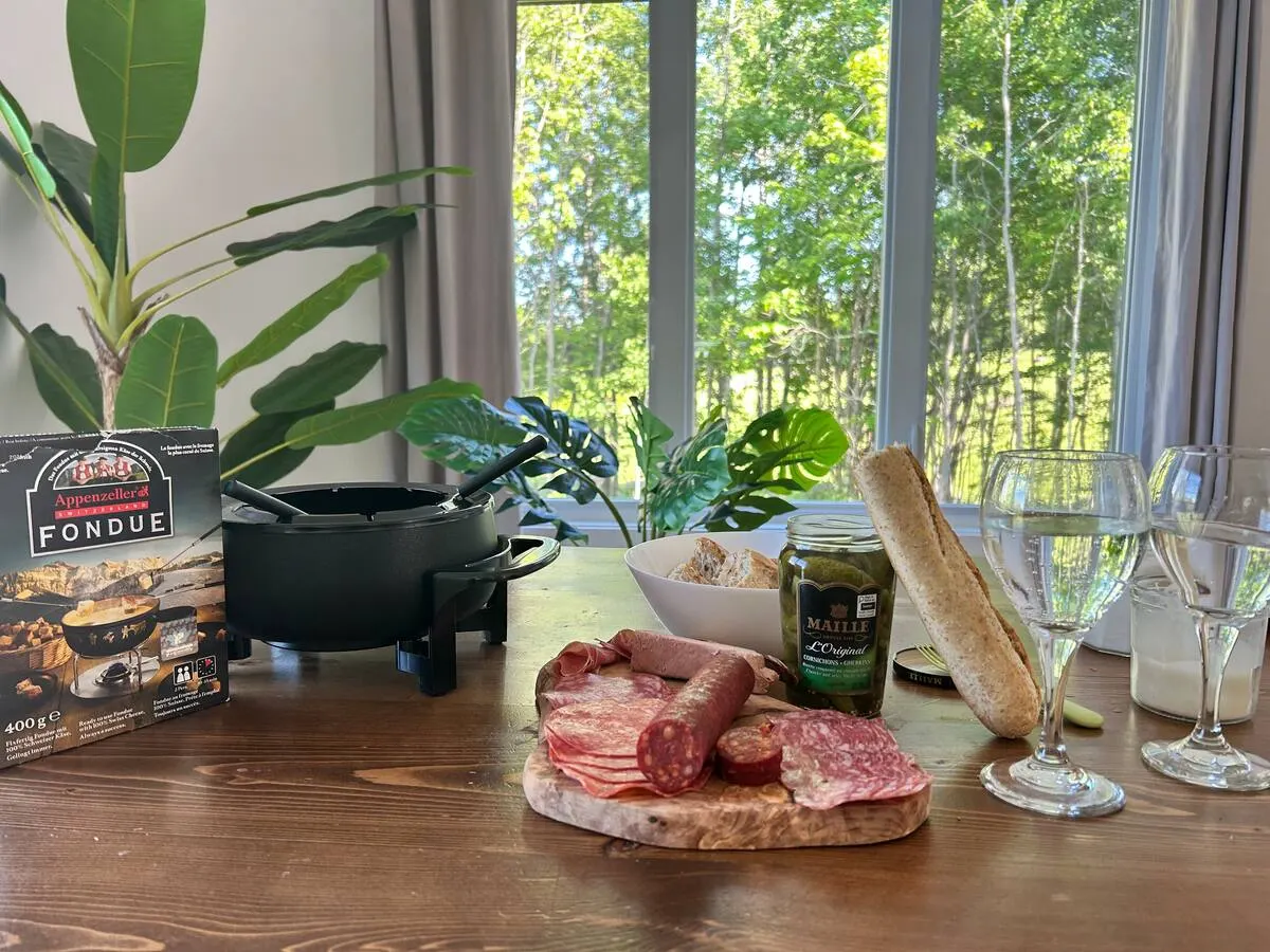 A fondue set, charcuterie, a baguette and wine glasses on a table by a window